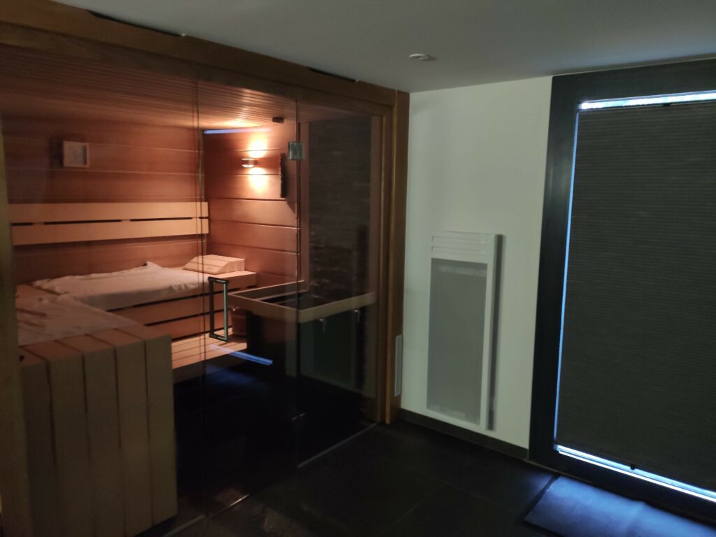Une cabine de sauna en bois éclairée à l'intérieur, visible à travers une paroi en verre dans une pièce moderne au sol sombre et aux murs blancs à Thonon-les-Bains