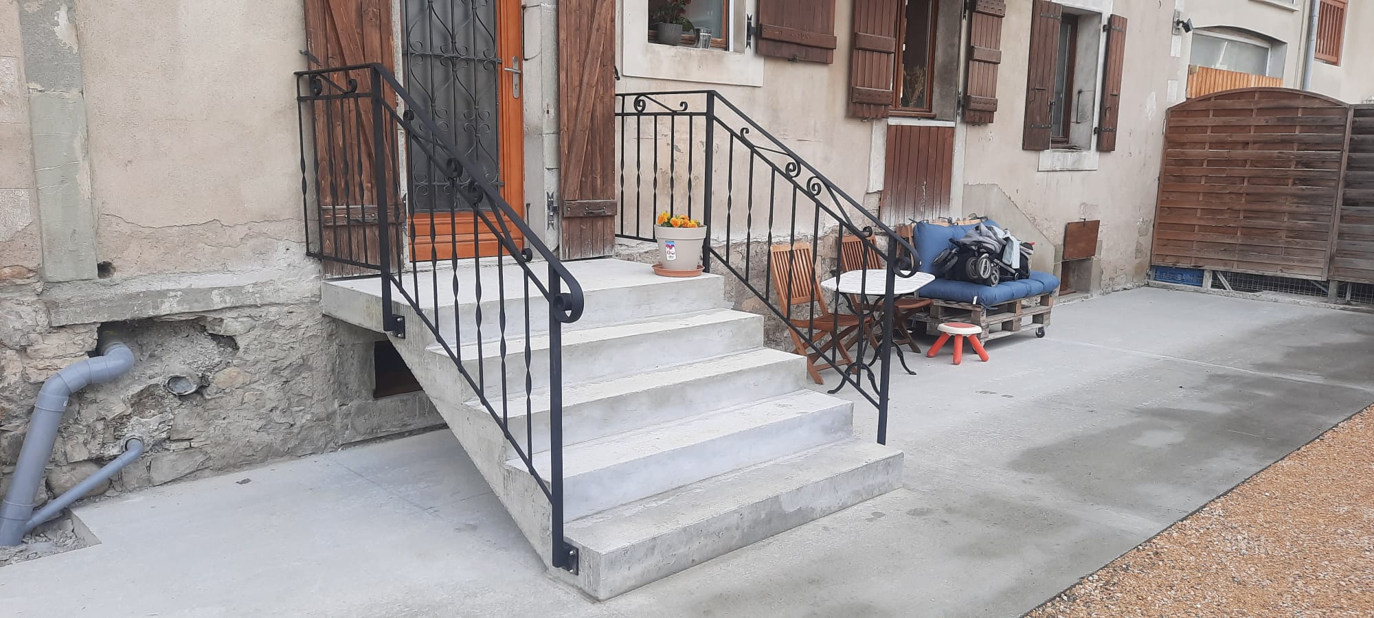 Escalier en béton avec rambarde en fer forgé menant à la porte d’entrée d’une maison ancienne rénovée. Terrasse aménagée avec table, chaises et banc en palettes.
