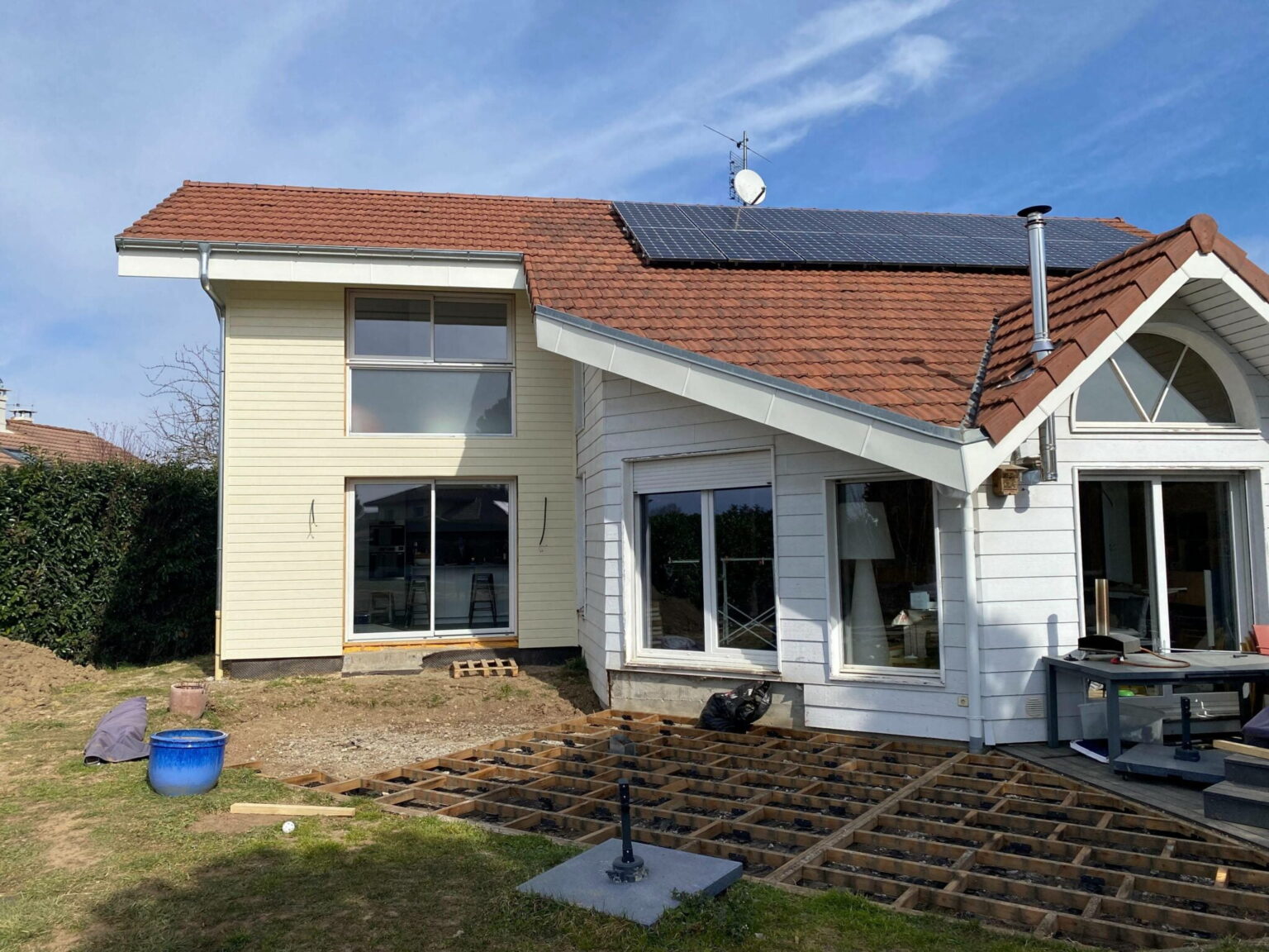 Maison en bois blanc avec toit en tuiles rouges, équipée de panneaux solaires et d’une terrasse en construction au premier plan.