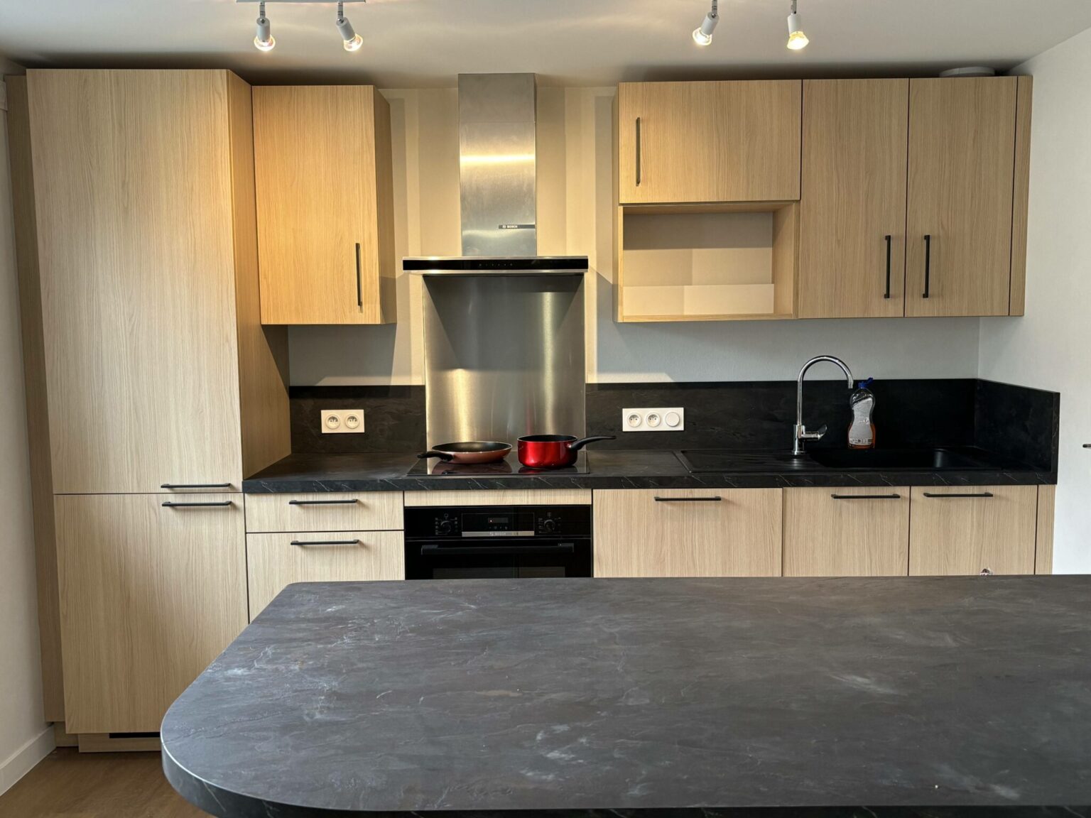 Cuisine moderne avec meubles en bois clair, plan de travail en pierre noire, hotte en acier inoxydable et îlot central.