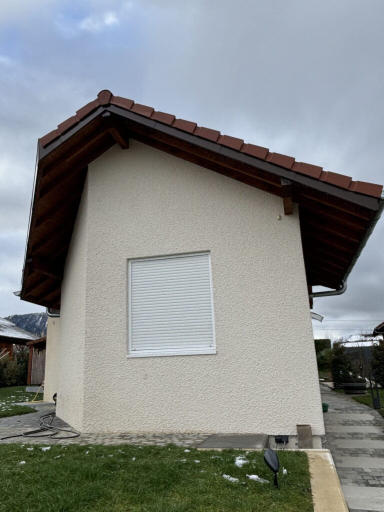 Vue latérale d’une maison crépie en beige, dotée d’un volet roulant blanc fermé et d’une charpente en bois sous un toit de tuiles.