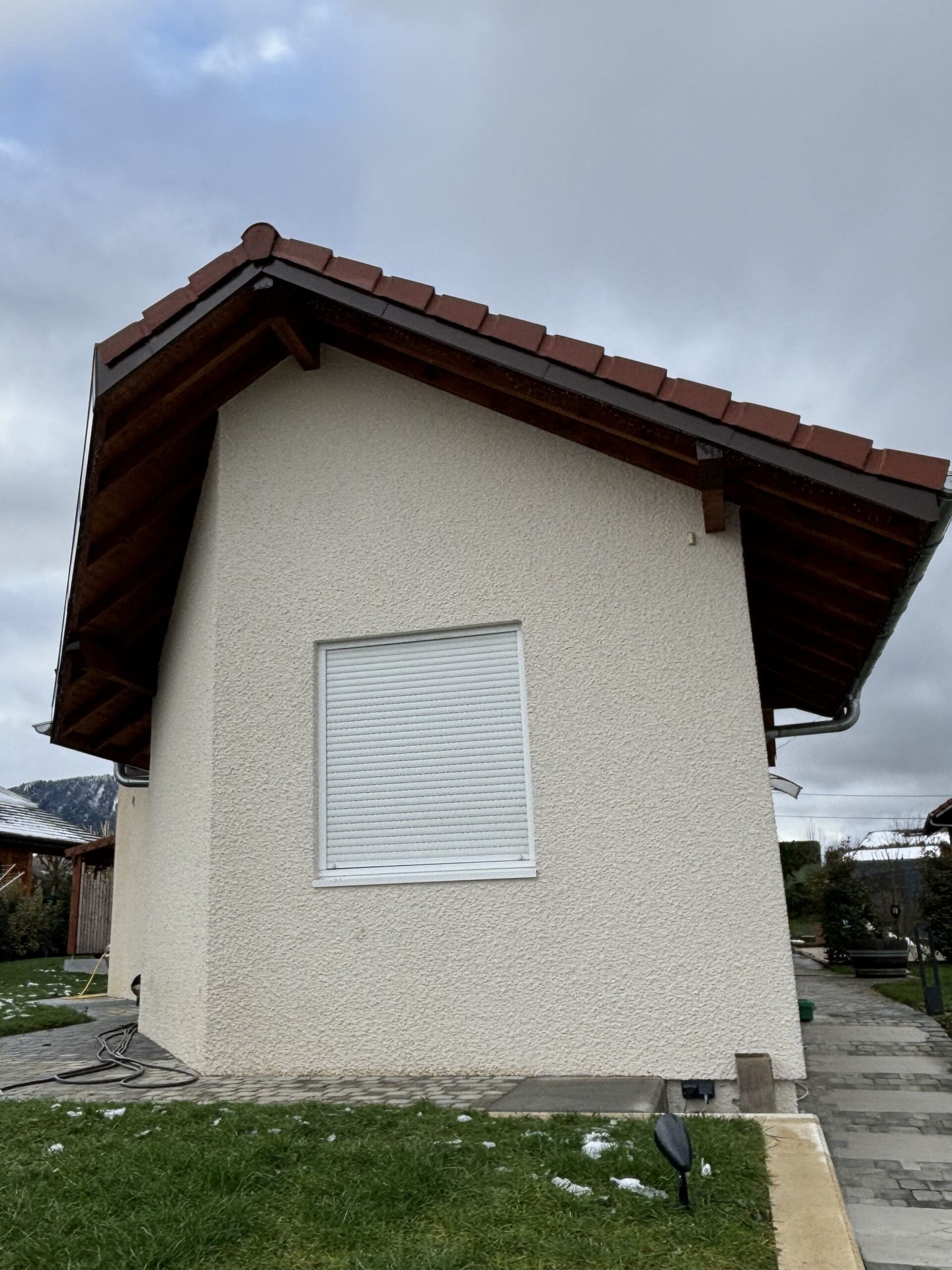 Vue latérale d’une maison crépie en beige, dotée d’un volet roulant blanc fermé et d’une charpente en bois sous un toit de tuiles.