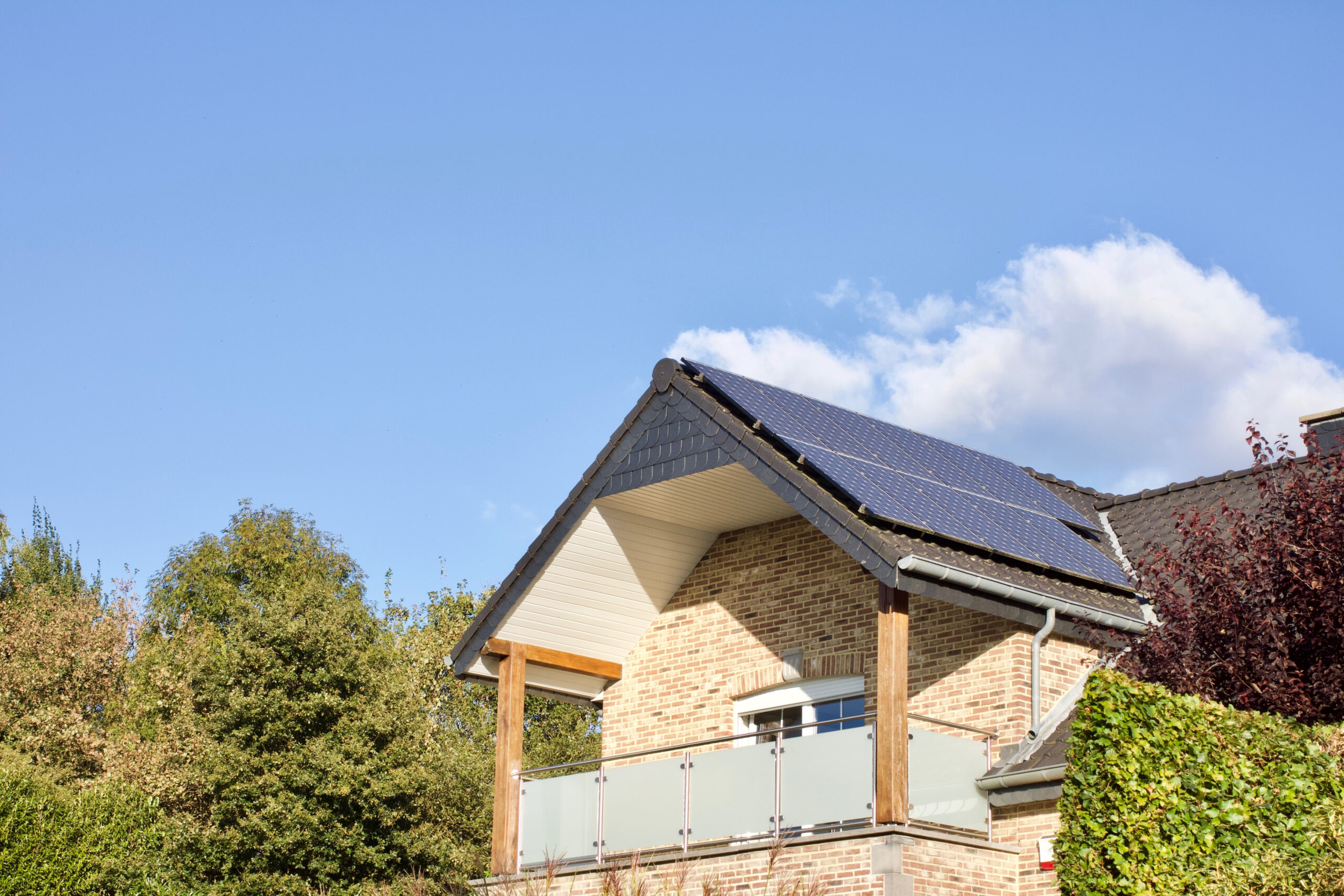 Maison en briques avec un toit équipé de panneaux solaires, un balcon en verre et une structure en bois, entourée de verdure sous un ciel dégagé.