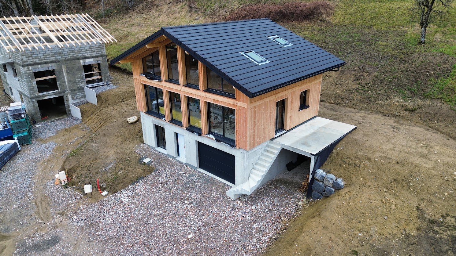 Vue aérienne d’une maison en ossature bois terminée, reposant sur un sous-sol en béton, avec une toiture en tuiles ardoise et de larges baies vitrées.