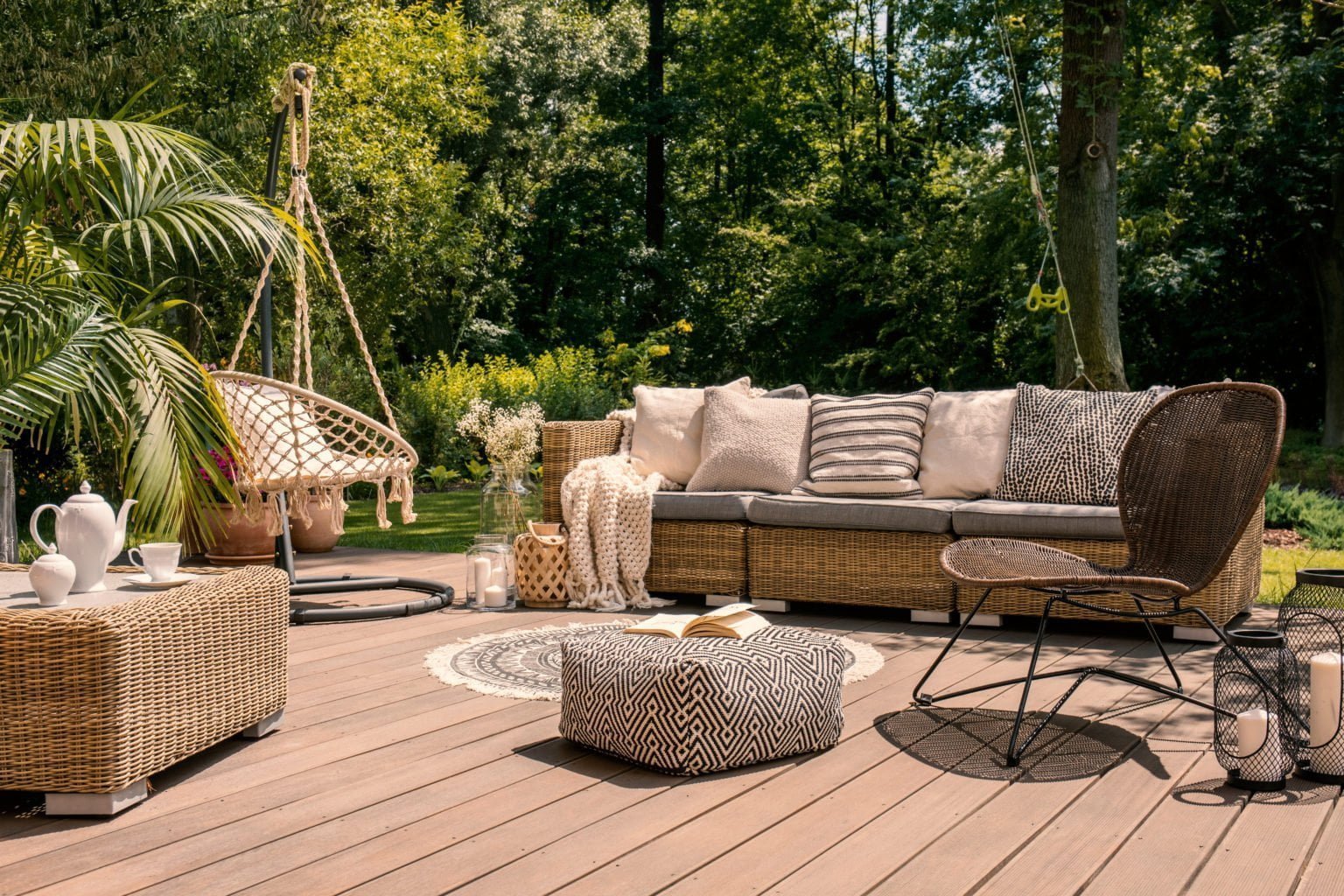 Un salon de jardin bohème chic avec un canapé en rotin, un fauteuil suspendu en macramé, une table basse en osier et un pouf à motifs, sur une terrasse en bois entourée de verdure.