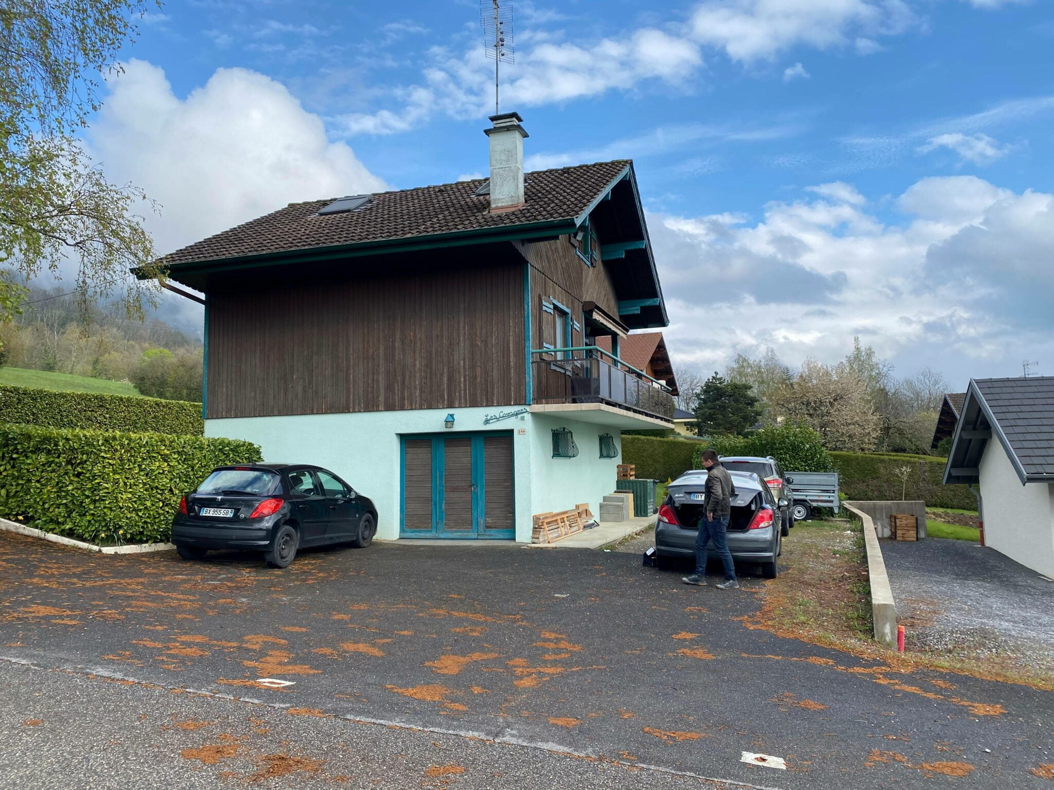 maison en bois avec voitures stationnées devant et personne chargeant le coffre
