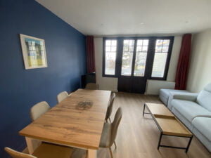 Salon rénové à Thonon avec parquet, mur bleu moderne, mobilier sobre et grandes fenêtres.
