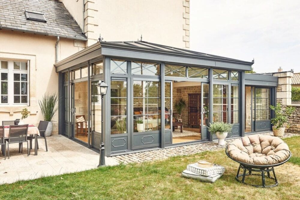 Extension de maison avec véranda vitrée et mobilier extérieur dans un jardin.
