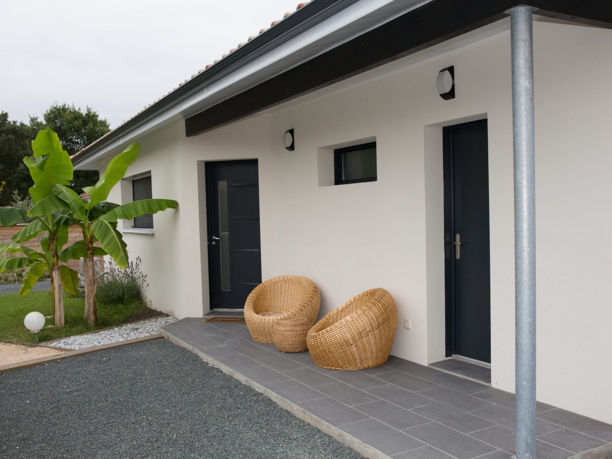Façade moderne rénovée avec un enduit clair, portes et fenêtres noires, terrasse en carrelage gris et mobilier extérieur en rotin.
