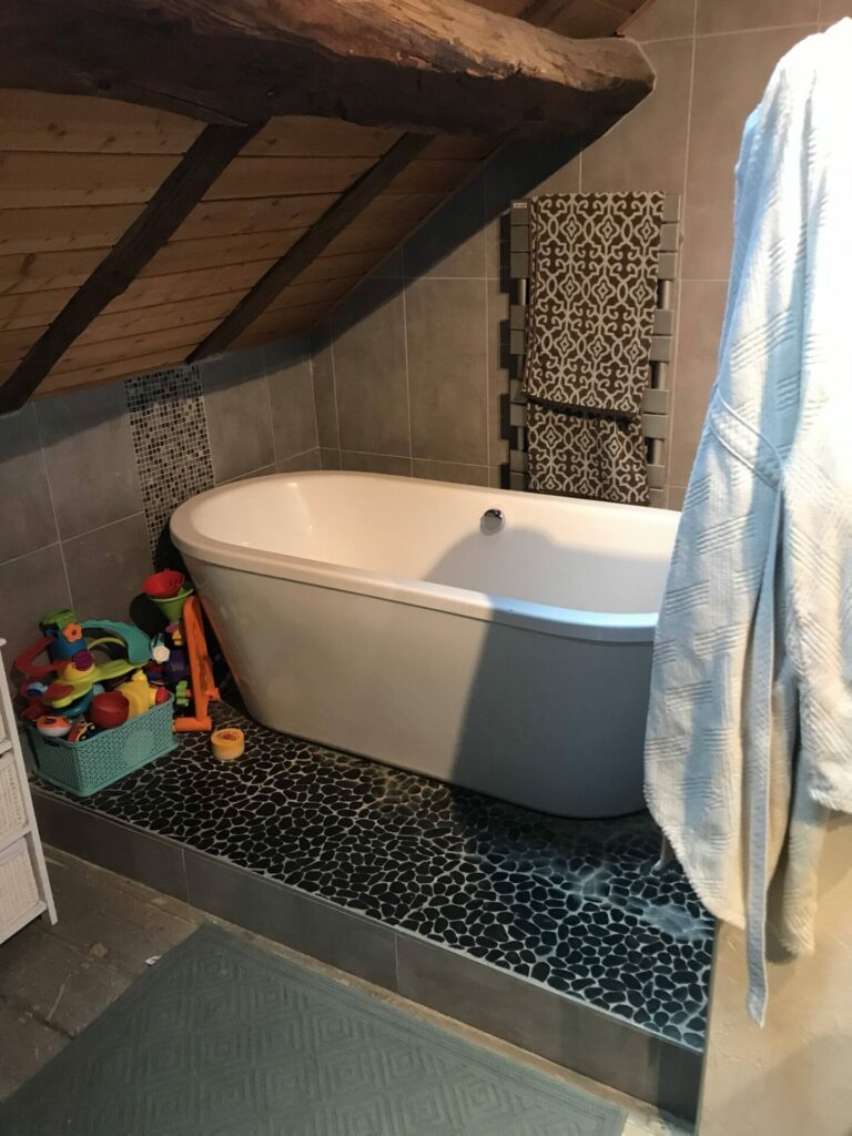 Baignoire îlot sous toiture en bois avec jouets d’enfants et carrelage effet galets.