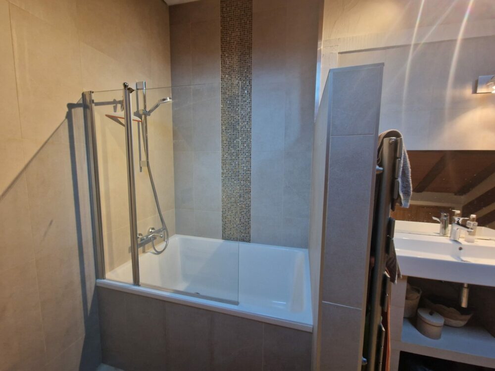 Baignoire encastrée avec carrelage beige et colonne de mosaïque dans une salle de bains rénovée.