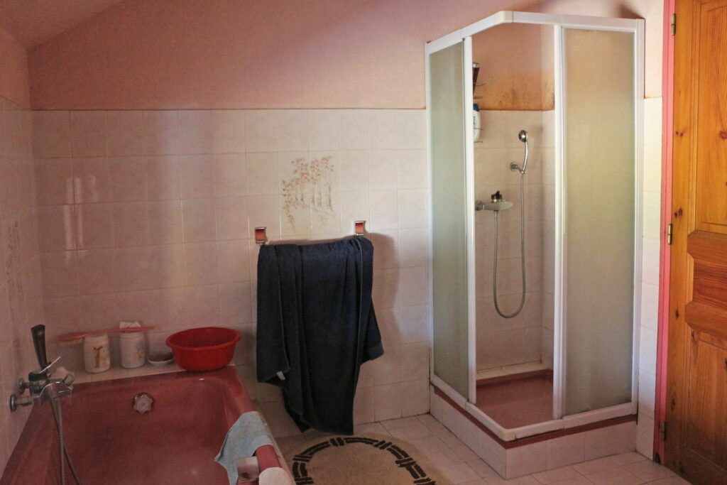 Ancienne salle de bain avec baignoire et douche rose sur fond de carrelage blanc.