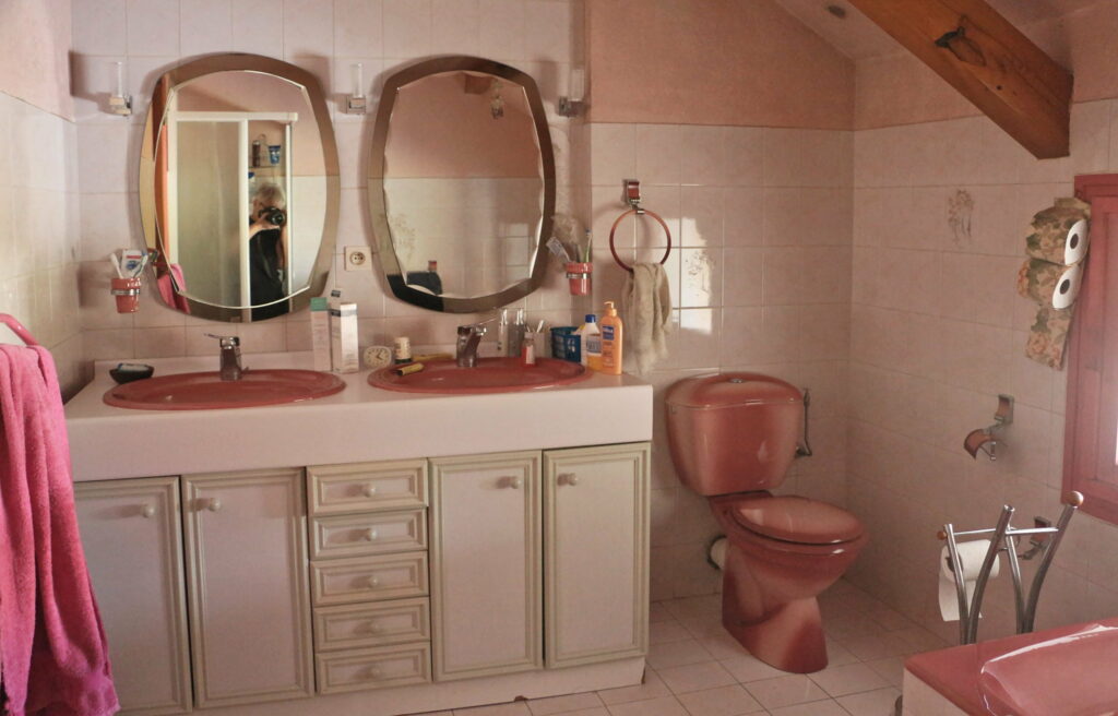 Ancienne double vasque rose avec meuble blanc et miroirs muraux ovales.