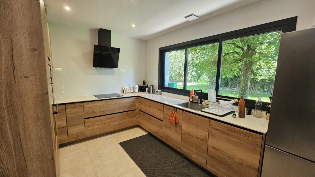 Cuisine moderne en L avec mobilier bois clair et grande fenêtre panoramique à Douvaine.