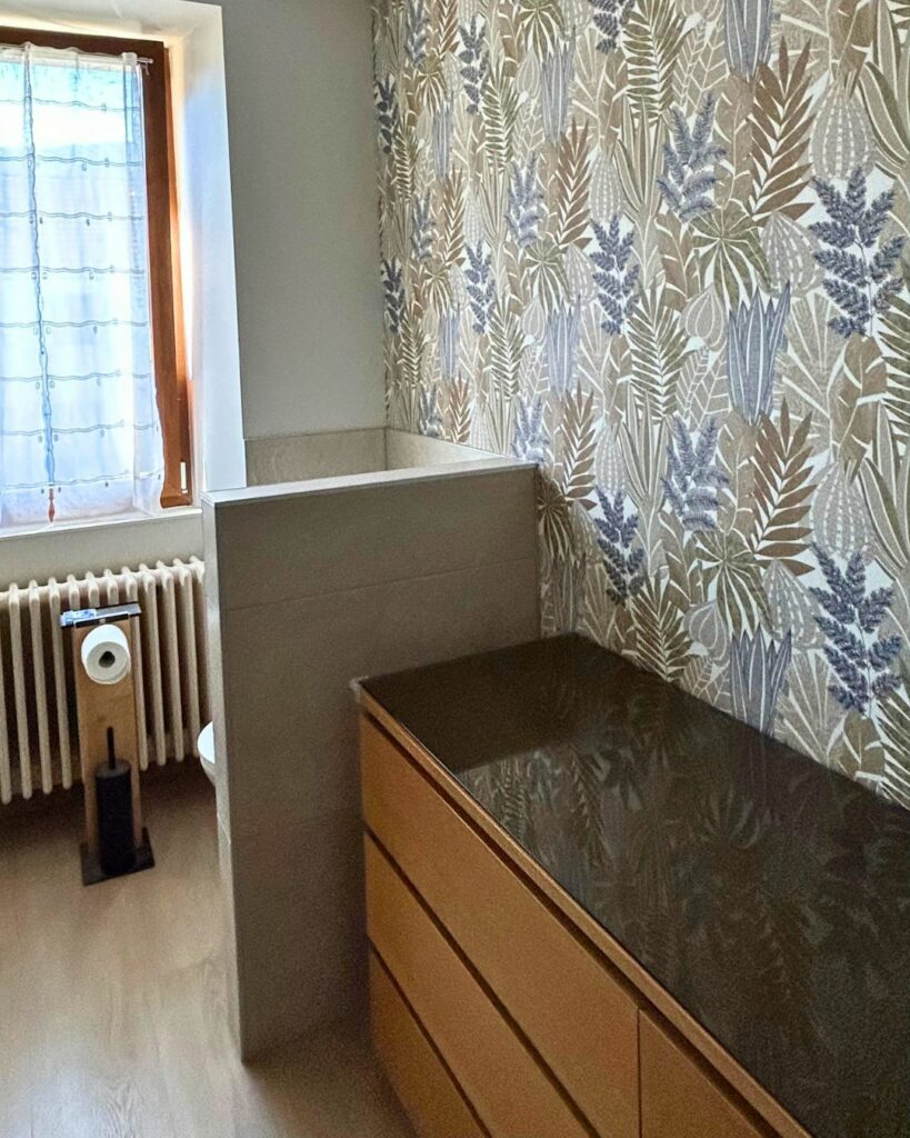 Vue latérale d’une salle de bain rénovée à Bons-en-Chablais avec meuble bois, plan vasque foncé et papier peint végétal.