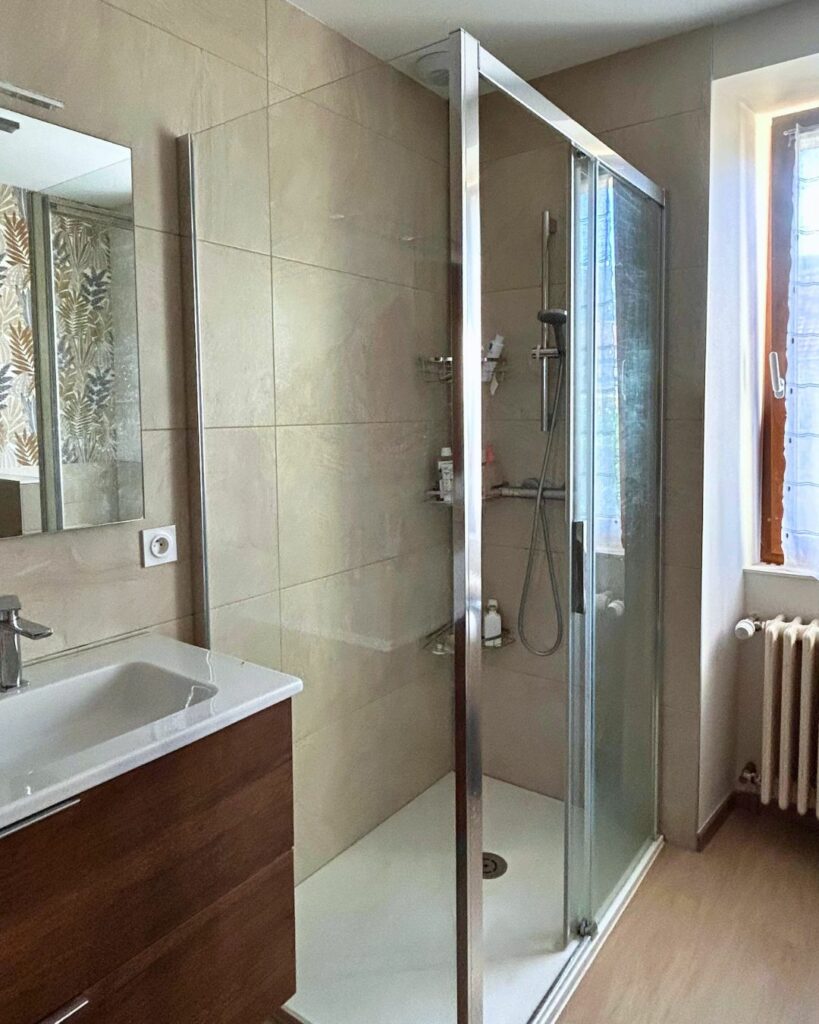 Salle de bain rénovée avec douche à l’italienne, carrelage beige, meuble vasque en bois foncé et lumière naturelle à Bons-en-Chablais.