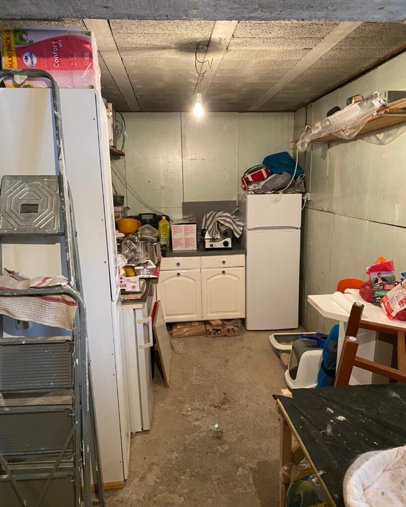 Garage encombré avant aménagement, sol brut et mur brut avec mobilier de fortune.