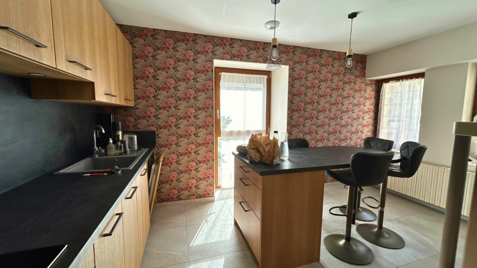 Cuisine rénovée d’une maison ancienne à Bons-en-Chablais avec papier peint décoratif, plan de travail noir mat et mobilier bois clair.