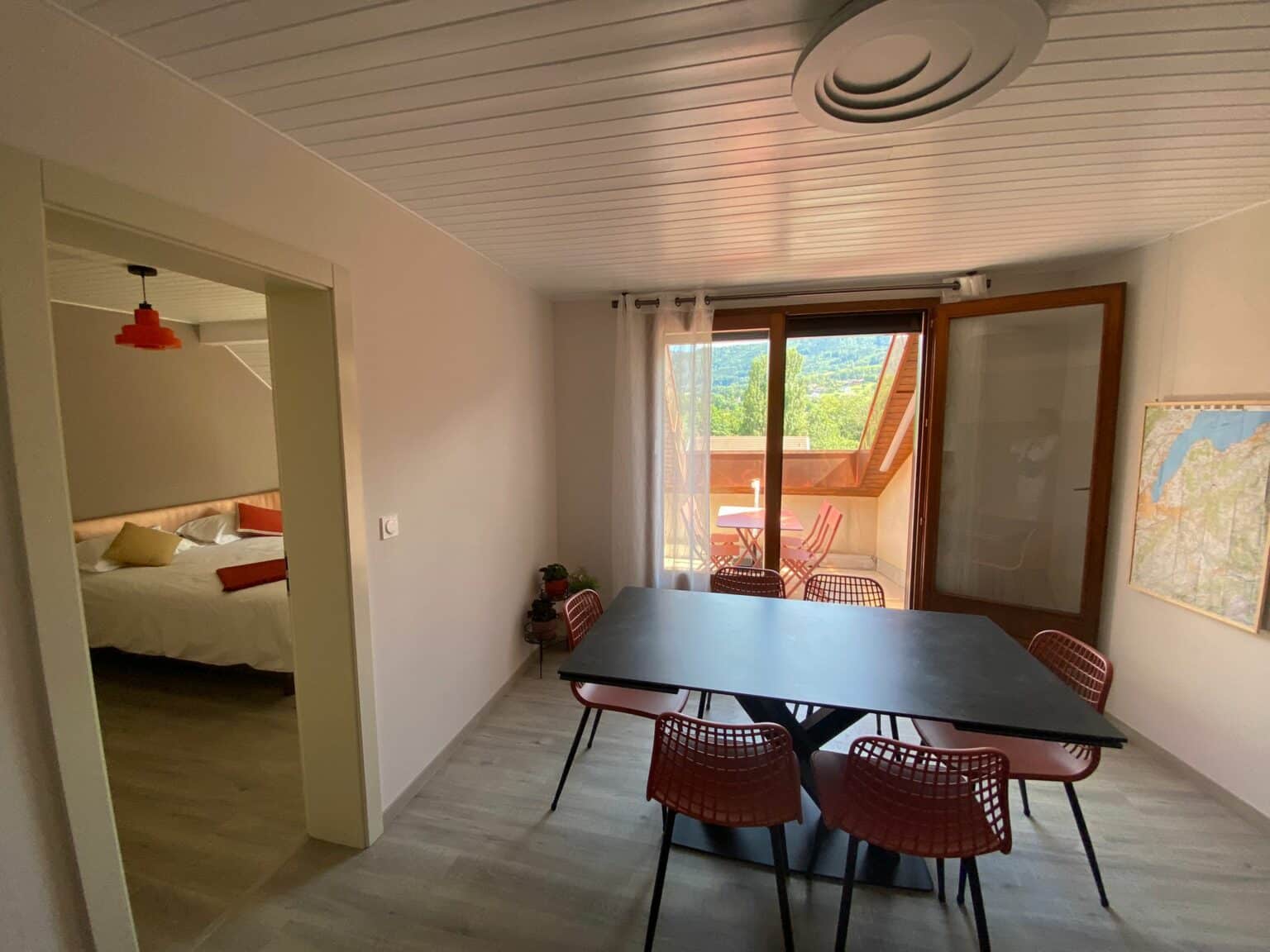 Salle à manger moderne sous les combles avec table noire, chaises rouges, accès à une terrasse avec vue montagne, et chambre double attenante visible sur la gauche.